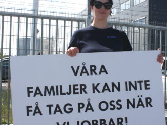 En person står utanför en stor lagerlokal med ett plakat ed texten "våra familjer kan inte få tag på oss när vi jobbar!"