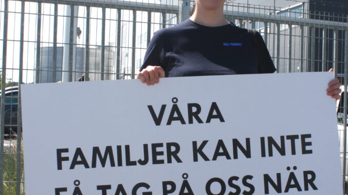 En person står utanför en stor lagerlokal med ett plakat ed texten "våra familjer kan inte få tag på oss när vi jobbar!"