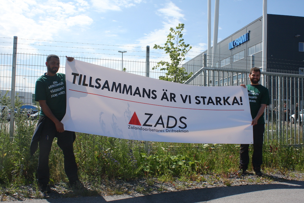 Två personer står utanför en lagerbyggnad och håller i en banderoll med texten "Tillsammans är vi starka, ZADS-Zalandoarbetares driftsektion"