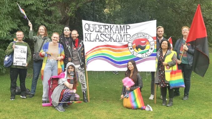 Tio personer är samlade vid en banderoll med texten Queerkamp, klasskamp. Samlingen har också regnbågsflaggor och rödsvarta flaggor.