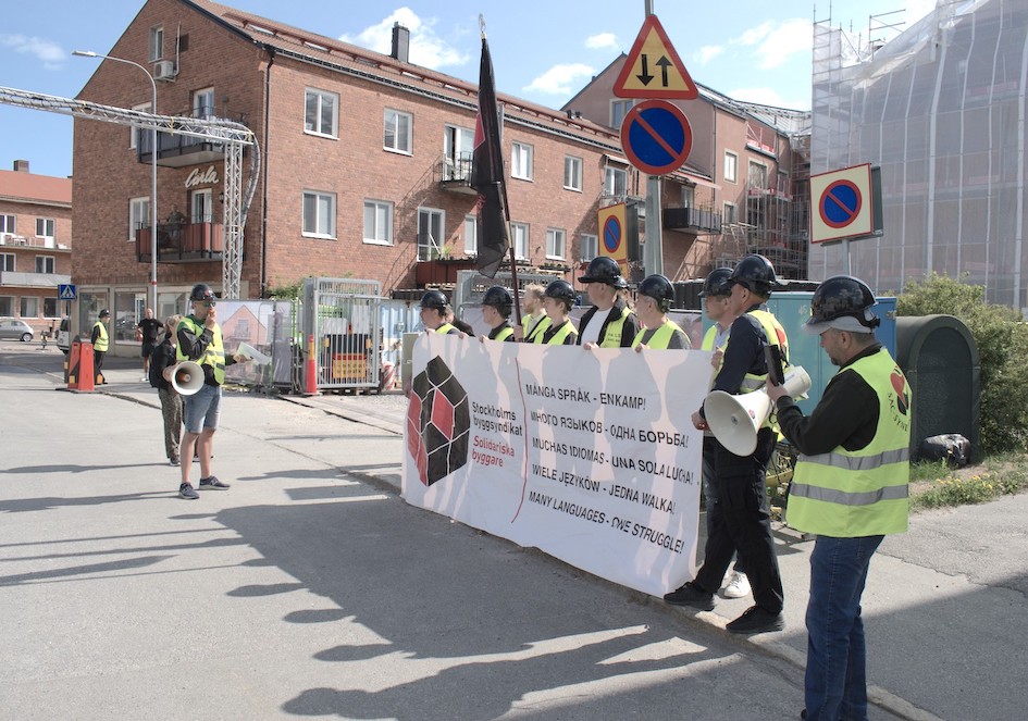 Utanför en byggarbetsplats står en fackförening som protesterar mot att arbetare på bygget inte fått den lön hen utlovats.