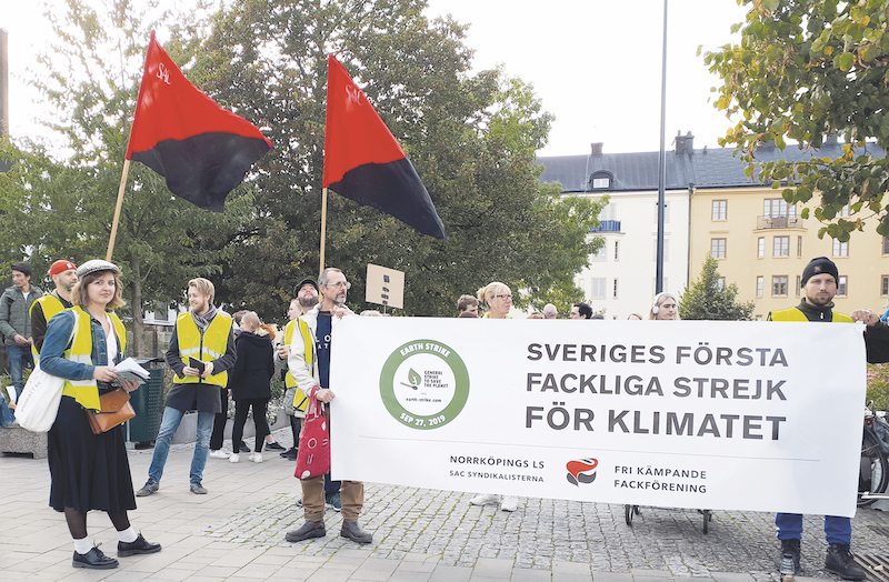 Människor på ett torg deltar i en demonstration. Två personer håller upp en banderoll med texten "Sveriges första fackliga strejk för klimatet, Norrköpings LS av SAC Syndikalisterna, fri kämpande fackförening". Två andra personer håller i rödsvarta fanor.