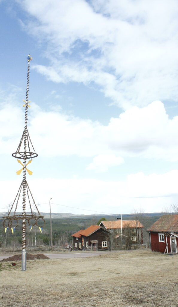 Vy i Skattungbyn, Orsa kommun. En kal midsommarstång och några röda stugor med kullar och himmel med moln i bakgrunden.