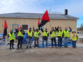 En samling människor med gula västar och rödsvarta fanor står på rad framför en byggarbetsplats.