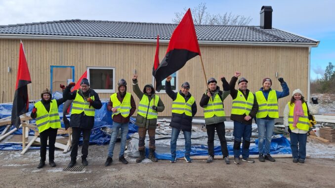 En samling människor med gula västar och rödsvarta fanor står på rad framför en byggarbetsplats.